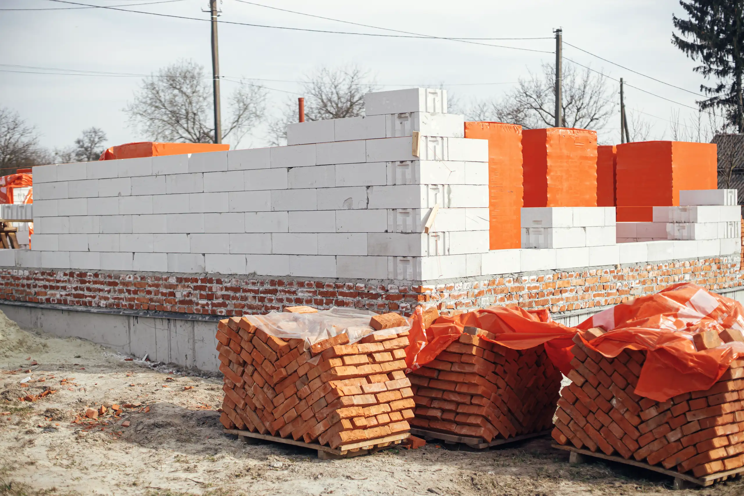 Ściany nośne z białych bloczków na świeżych fundamentach, obok stosy cegieł gotowe do murowania